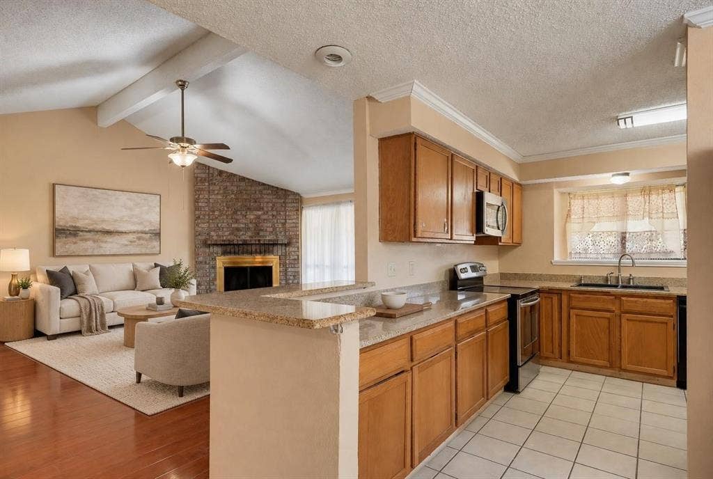 Kitchen featuring a breakfast bar with stone-look countertop, tile flooring, and wood cabinetry, paired with a living area showcasing a vaulted ceiling, exposed brick fireplace, and wood-finish flooring