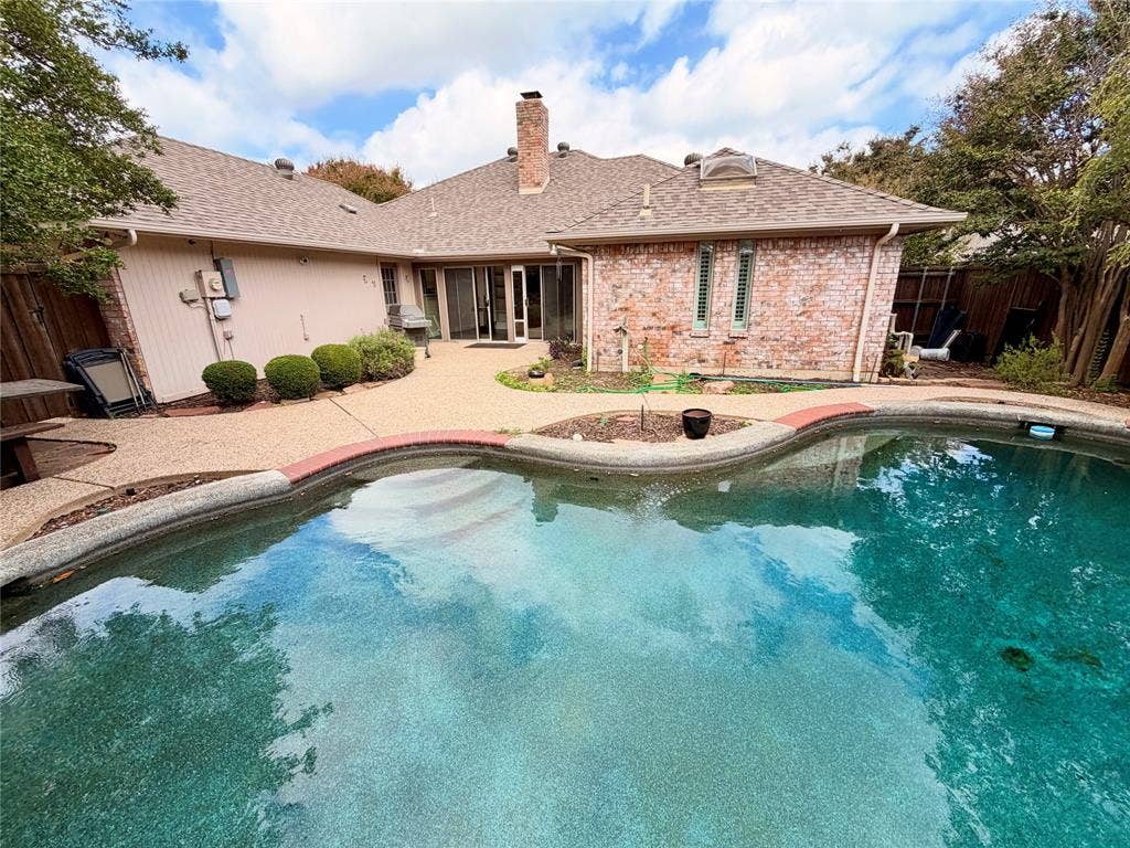 Backyard featuring a custom-shaped swimming pool with brick coping, an expansive aggregate patio, a prominent brick chimney, and multi-pane glass sliding doors