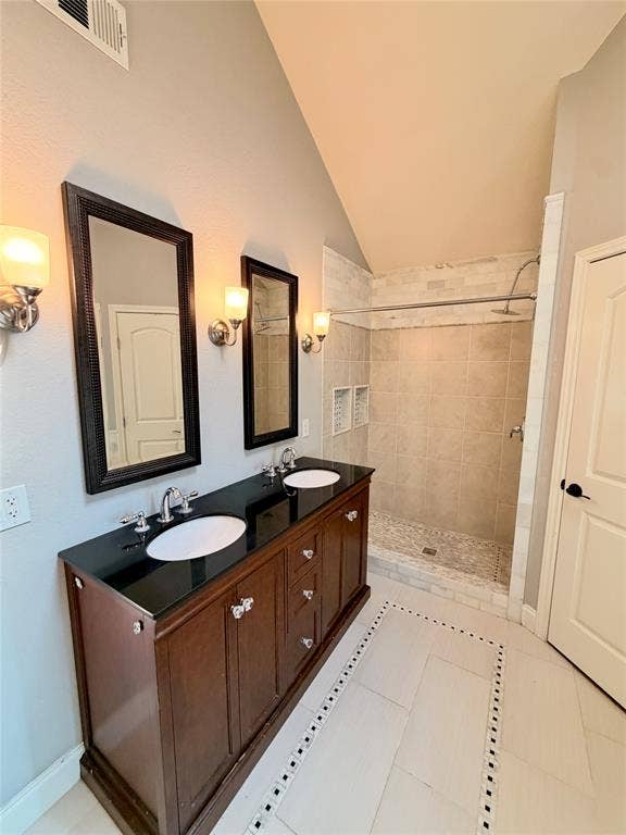 Dual-sink vanity with dark stone countertop, two framed mirrors, wall-mounted light fixtures, vaulted ceiling, and walk-in shower with tiled surround and built-in niches