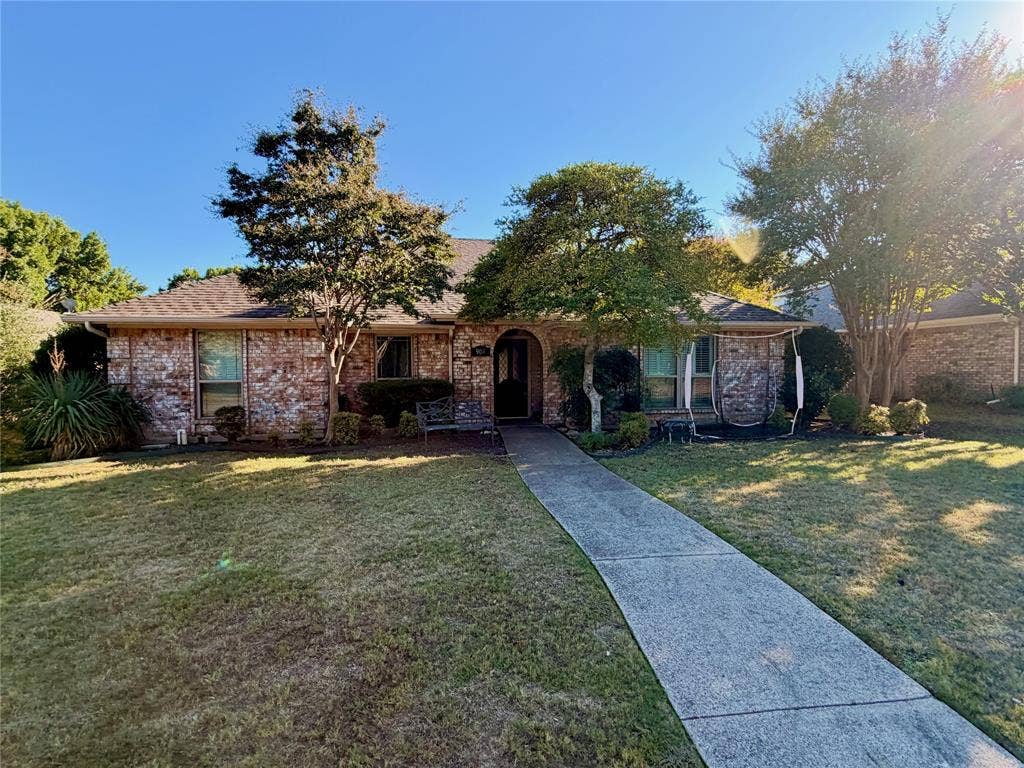 Single-story brick exterior with an arched entryway, shingled roof, concrete walkway, and mature trees