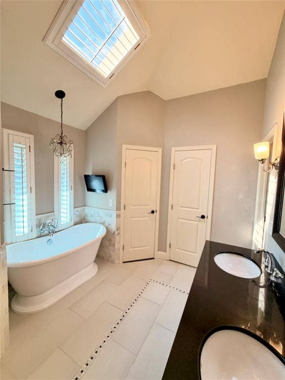Vaulted ceiling featuring a skylight, freestanding soaking tub, dual-sink vanity with dark stone countertop, and two interior doors with arched panels