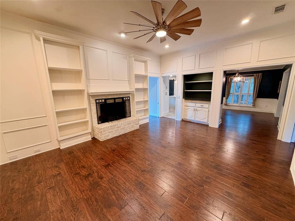 Living area featuring wood-finish flooring, wainscoting, a brick fireplace with built-in shelving, and a ceiling fan with a multi-blade design