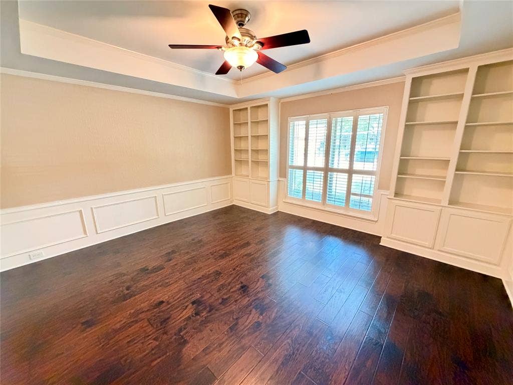 Room featuring wood-finish flooring, tray ceiling with a central ceiling fan, white wainscoting, and floor-to-ceiling built-in cabinetry