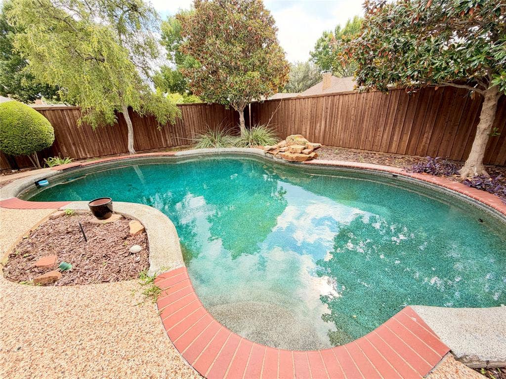 Curved swimming pool featuring a decorative rock water feature, red brick coping, textured concrete pool deck, and perimeter wood fencing