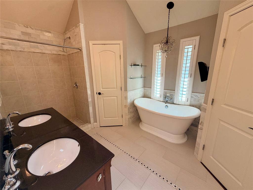 Double vanity with dark countertops and integrated oval sinks, freestanding soaking tub, walk-in tile shower, tile flooring, and vaulted ceiling