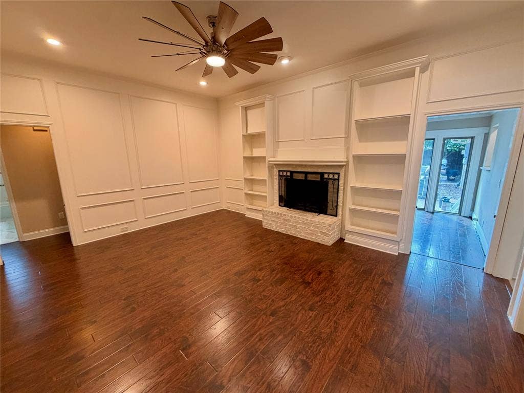 Wood-finish flooring, fireplace with a textured masonry hearth, floor-to-ceiling built-in shelving, wall molding, and a multi-blade ceiling fan