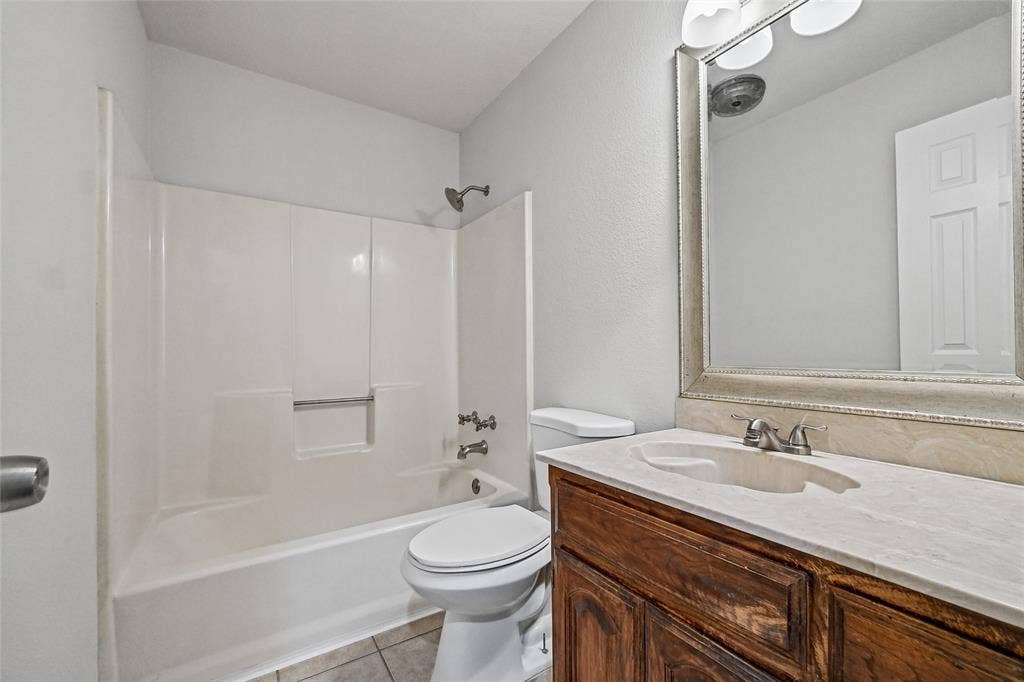 7929 Sunrise Drive, Watauga, TX 76148 Bathroom featuring a wood-finish vanity with a stone-surface countertop, an integrated sink with a dual-handle faucet, a framed mirror with integrated lighting, and a tub-shower combination with a molded surround