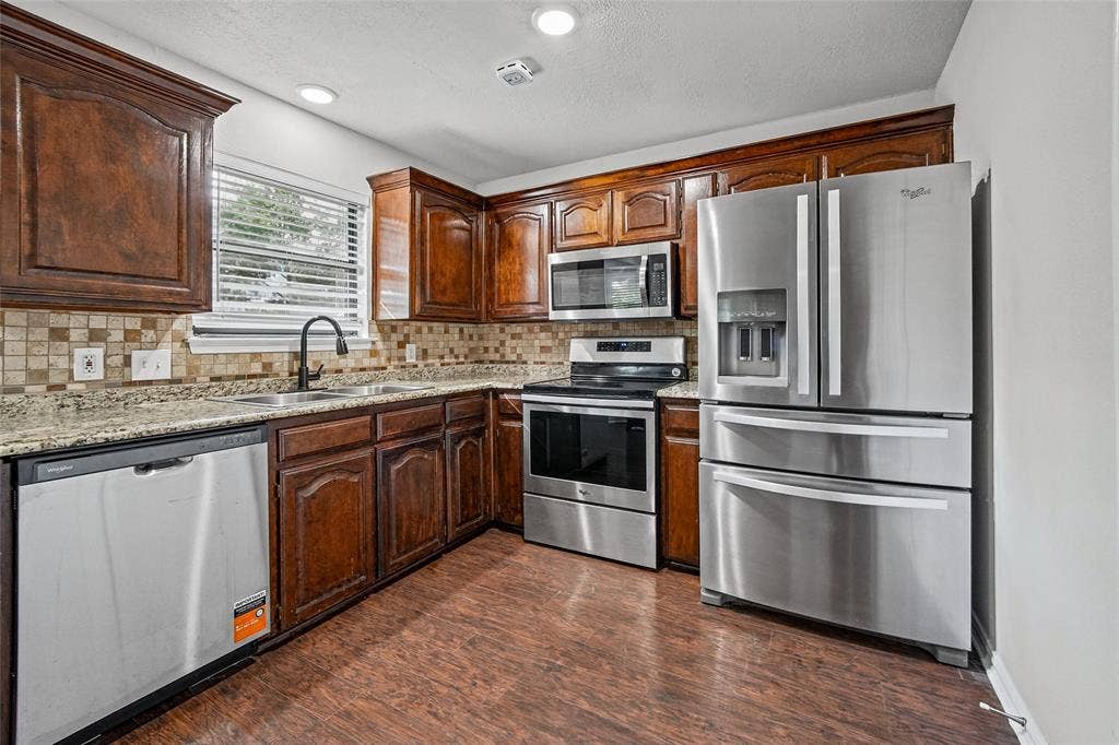 7929 Sunrise Drive, Watauga, TX 76148 Kitchen featuring dark wood cabinetry, stone-surface countertops, a tiled backsplash, wood-finish flooring, and stainless steel appliances