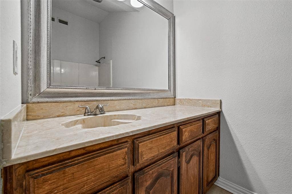 7929 Sunrise Drive, Watauga, TX 76148 Vanity featuring natural stone-finish countertop, integrated sink, dual-handle faucet, and wood-finish cabinetry topped with a framed mirror