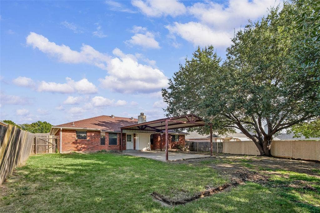 7929 Sunrise Drive, Watauga, TX 76148 Rear exterior featuring a brick facade, covered patio area with metal supports, fenced yard, and a mature tree