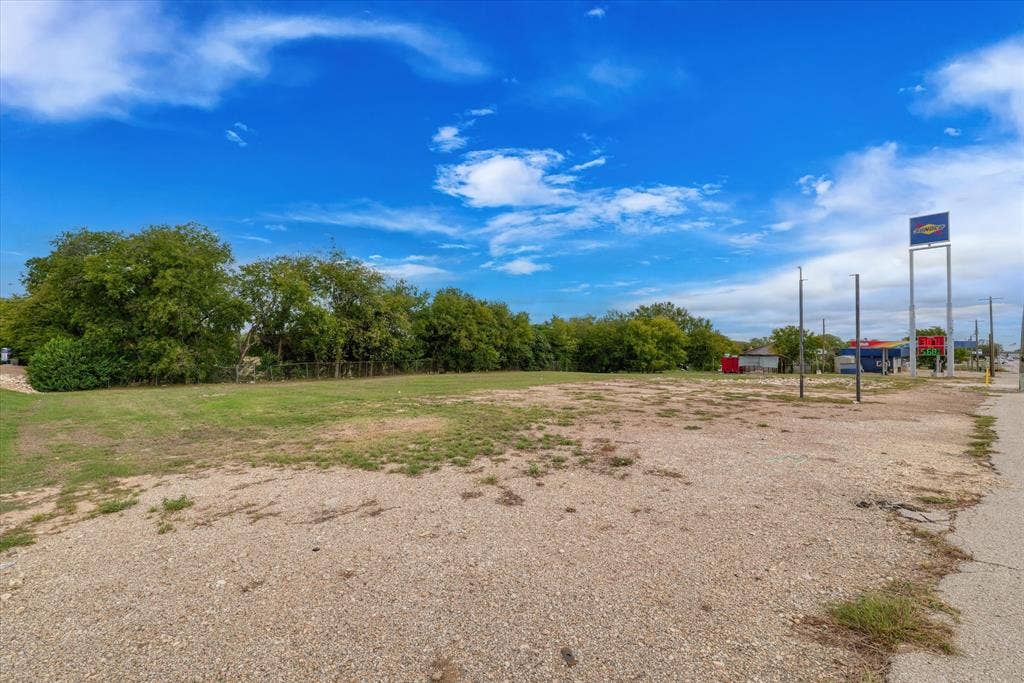 Expansive open lot featuring a combination of cleared gravel surfacing and grassy areas, delineated by mature perimeter trees and chain-link fencing