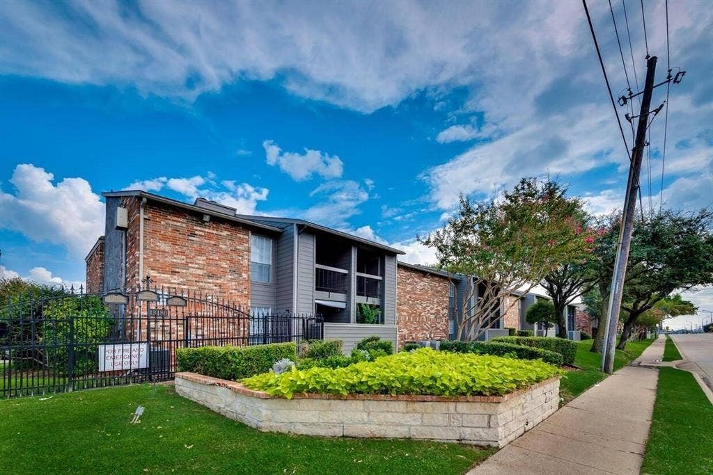 12484 Abrams Road, Dallas, TX 75243 Multi-story building exterior featuring red brick and dark-toned paneling, raised stone-block landscaping, wrought-iron perimeter fencing, and mature trees