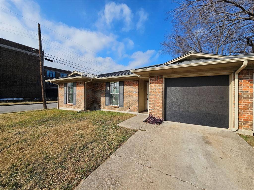 Single-story brick exterior featuring a metal roof, attached garage, concrete driveway, and shutters
