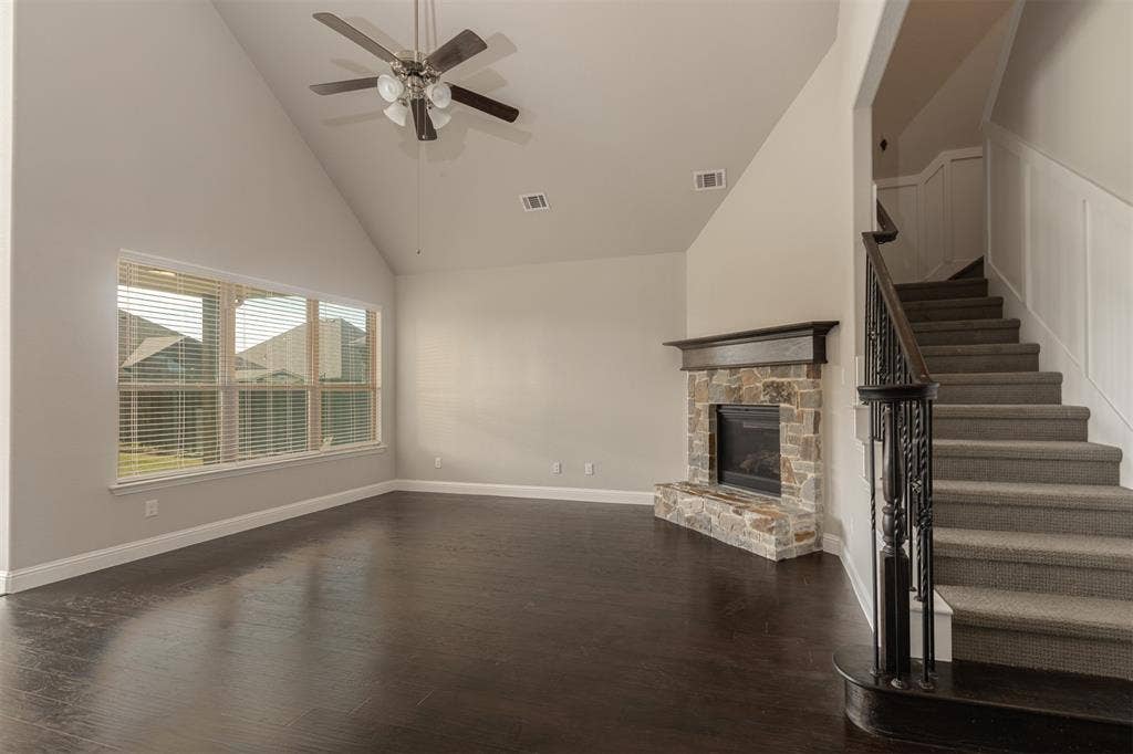 Vaulted ceiling featuring a ceiling fan, stone-surround fireplace with a wooden mantel, wood-finish flooring, and a staircase with a decorative iron balustrade