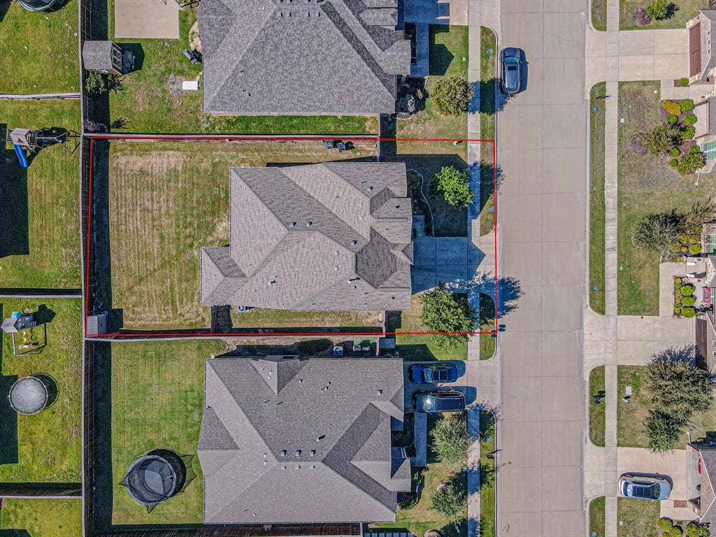 Aerial perspective of a residential lot featuring a concrete driveway, perimeter fencing, and a multi-pitched shingled roof