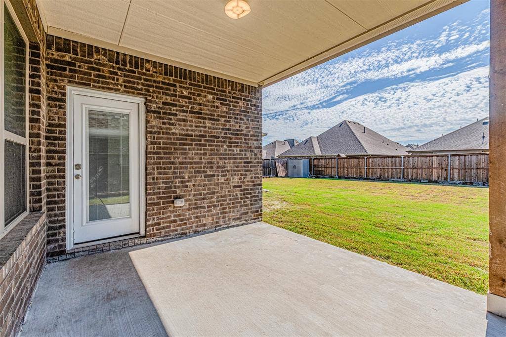 Covered concrete patio featuring a brick exterior wall, full-light glass door, and integrated ceiling lighting, opening onto an expansive fenced backyard