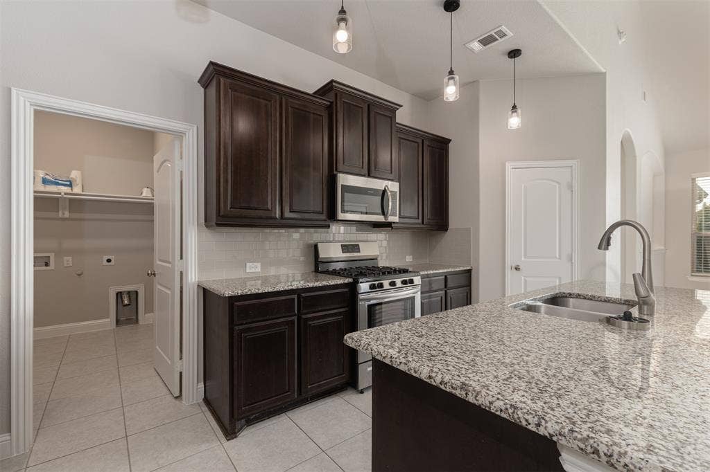 Kitchen featuring dark wood cabinetry, stone countertops, stainless steel appliances, tile backsplash, and pendant lighting