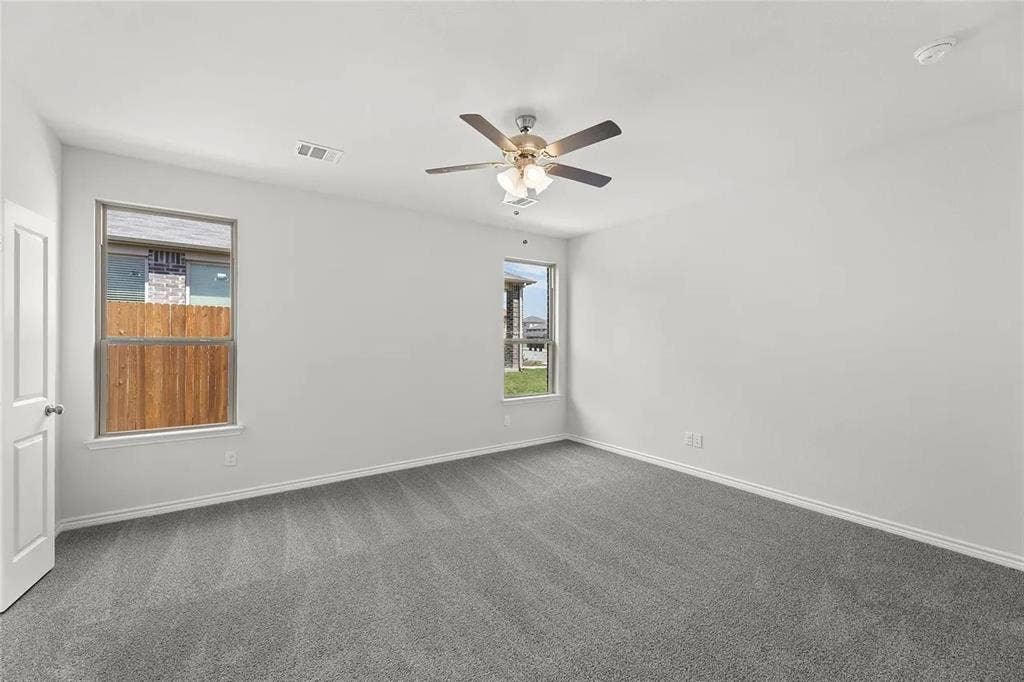 Neutral-toned interior room featuring wall-to-wall carpeting, dual windows with exterior views, and a multi-blade ceiling fan with integrated lighting