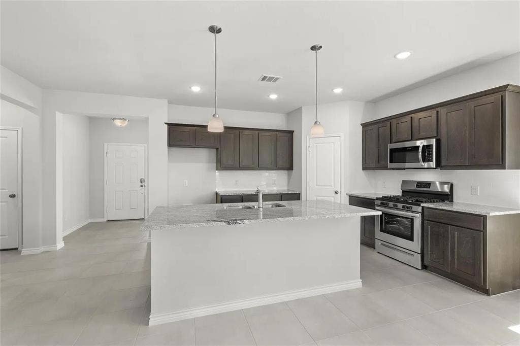 Kitchen featuring dark-toned cabinetry, stone-finish countertops, stainless steel appliances, a central island with an integrated sink, and dual overhead pendant lighting