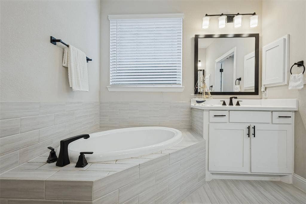 5313 Pratt Circle, McKinney, TX 75071 Soaking tub surrounded by horizontal tile, white vanity cabinet with a solid-surface countertop, matte black fixtures, framed vanity mirror, and linear vanity lighting