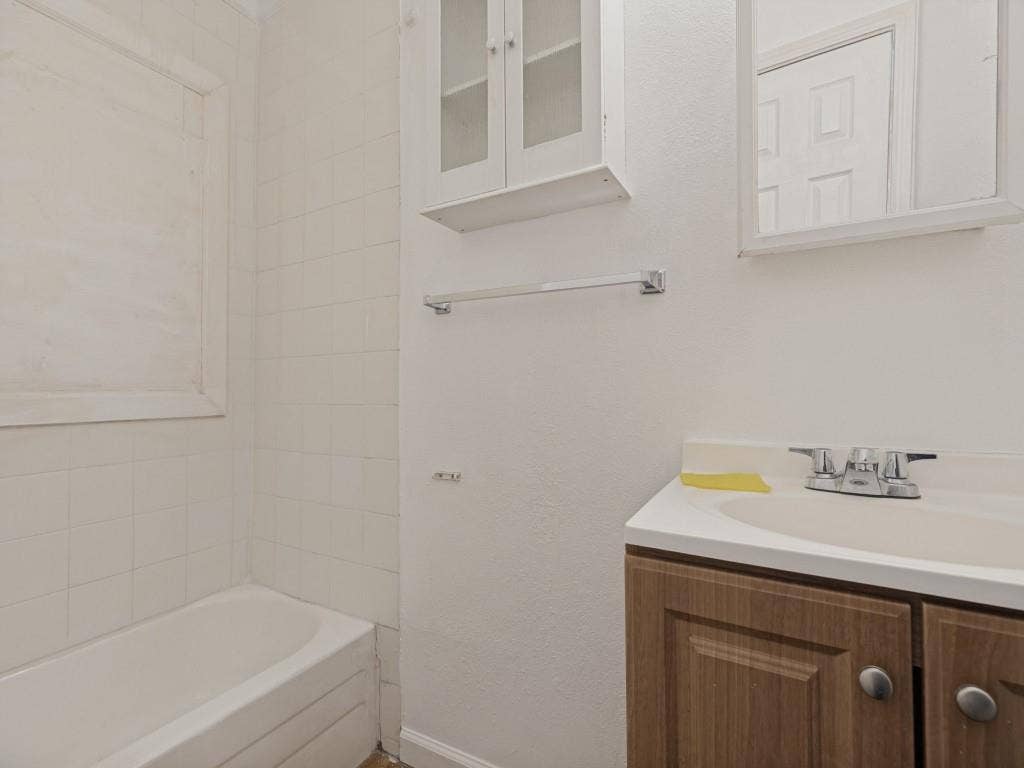 2009 Pecan Street, Commerce, TX 75428 Bathroom featuring a wood-finish vanity with integrated sink, white tile tub surround, wall-mounted cabinet with glass doors, and a medicine cabinet with mirror
