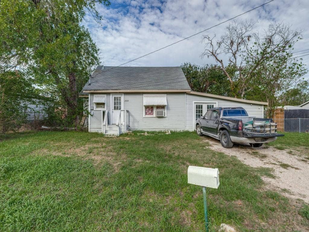2009 Pecan Street, Commerce, TX 75428 Single-story exterior featuring light gray horizontal siding, an asphalt shingle roof, glass-panel entry door, and a gravel driveway