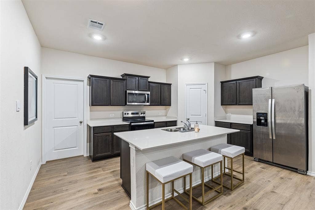 Kitchen featuring dark cabinetry, quartz countertops, a central island with an integrated stainless steel sink, stainless steel appliances, and wood-finish flooring