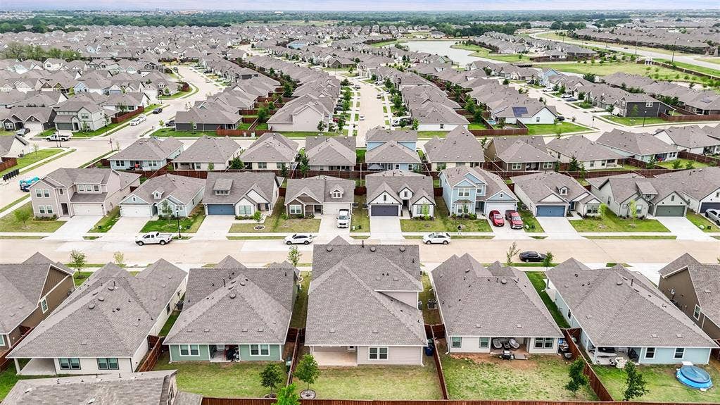Aerial perspective of a residential neighborhood featuring uniform single-story homes with pitched roofs, paved streets, and wood-fencing property borders