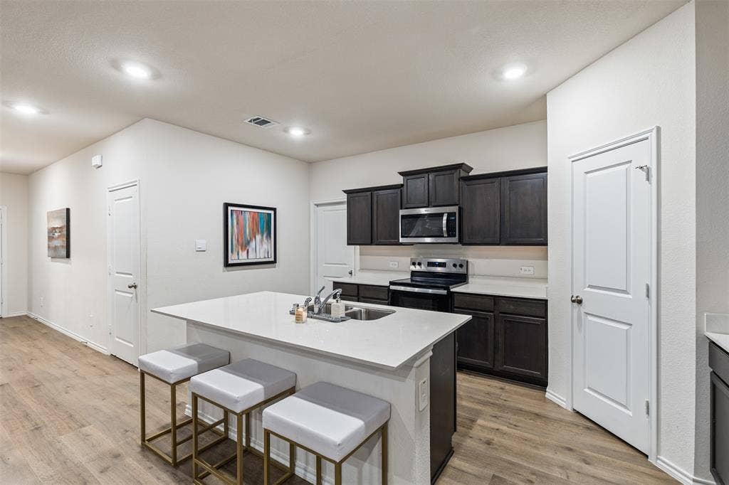 Kitchen featuring dark-finish cabinetry, white solid-surface countertops, a center island with an integrated stainless steel dual-basin sink, recessed ceiling lighting, and wood-finish flooring