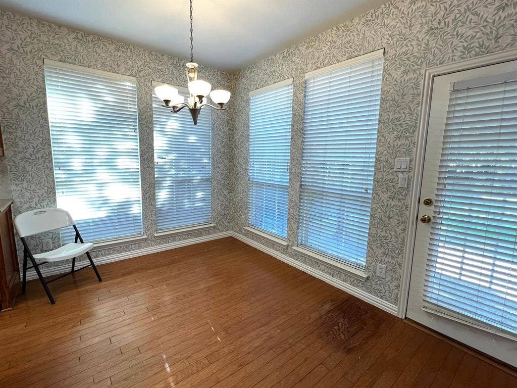 Hardwood flooring, patterned wallpaper, four large windows with horizontal blinds, multi-arm chandelier, and a glass-paneled exterior door