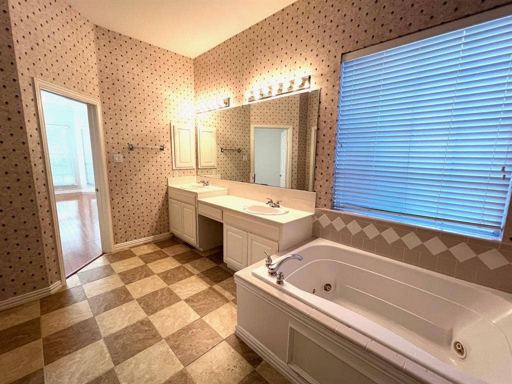 Dual-sink vanity with white cabinetry and solid-surface countertop, soaking tub with tile surround, and expansive wall-mounted mirror