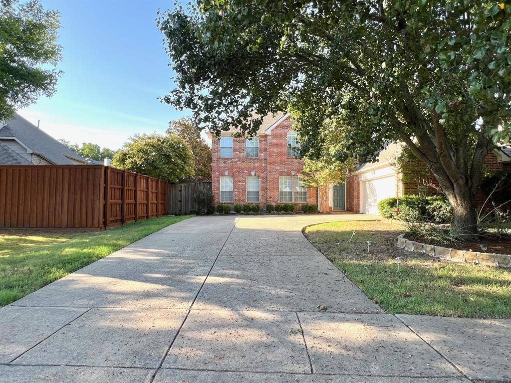 Two-story brick exterior featuring a multi-car garage, concrete driveway, wood-finish perimeter fencing, and established trees within the front lawn
