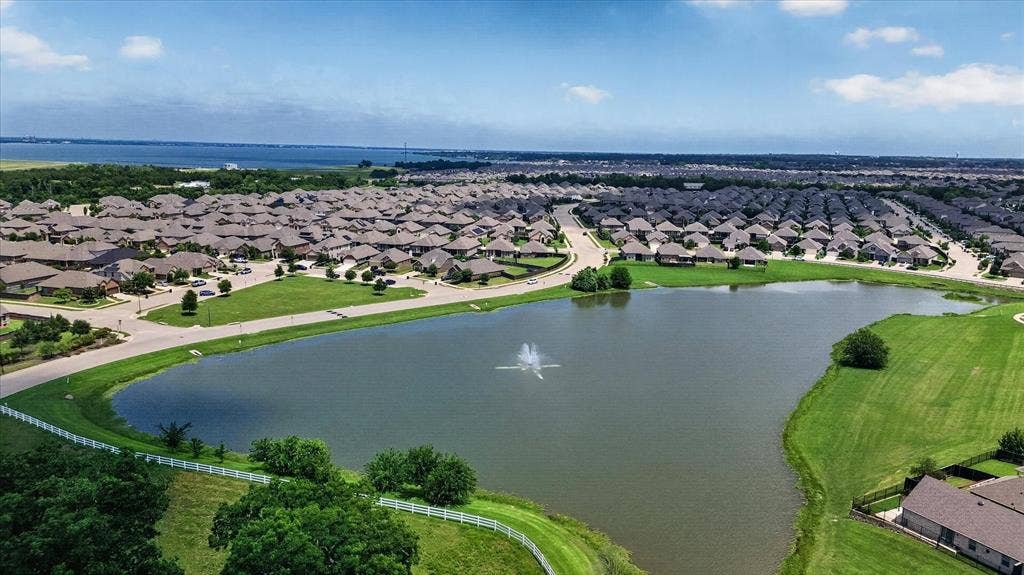 Aerial perspective of a community pond featuring a central water fountain, surrounding green space, and white perimeter fencing