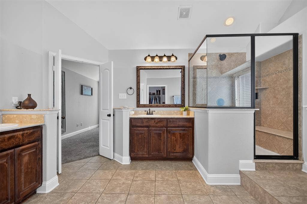122 Anns Way, Forney, TX 75126 Bathroom featuring dual dark wood vanities with stone countertops, a large framed mirror, and a glass-enclosed shower with tiled walls and a built-in bench