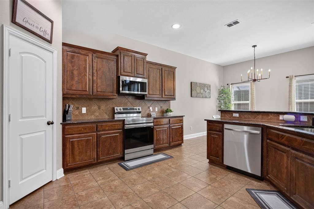 122 Anns Way, Forney, TX 75126 Kitchen featuring stained wood cabinetry, stone-tile backsplash, stainless steel appliances, dark countertops, and tile flooring