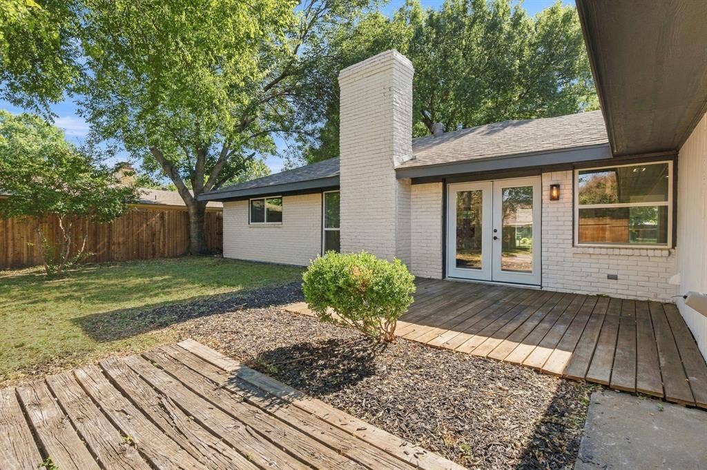 Exterior featuring white brick facade, masonry chimney, wood-plank decking, double glass-panel doors, and fenced backyard with mature trees