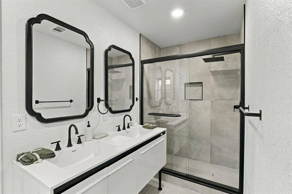 Floating dual-sink vanity with matte black faucets and hardware, expansive wall-mounted mirrors with black frames, and a walk-in shower featuring large-format tile surround and matte black fixtures