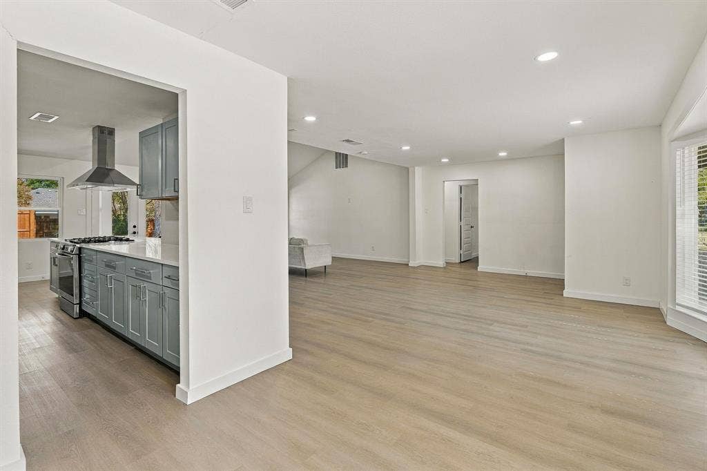 Open-concept living space featuring wood-finish flooring, recessed lighting, neutral wall paint, a bay window, and a kitchen equipped with shaker-style cabinetry, stone countertops, and a stainless steel vent hood