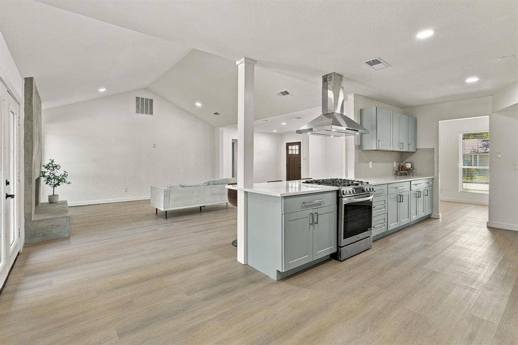 Vaulted ceilings, wood-finish flooring, kitchen island with stainless steel gas range, stainless steel vent hood, and light-toned shaker-style cabinetry