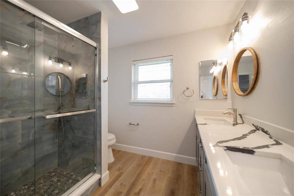 4333 Harvest Hill Road, Dallas, TX 75244 Bathroom featuring a walk-in shower with stone tile surround, double-sink vanity with patterned stone countertop, circular wood-frame mirrors, and wood-finish flooring