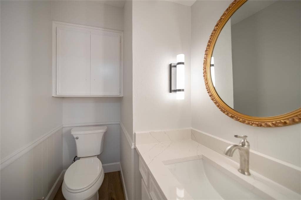 4333 Harvest Hill Road, Dallas, TX 75244 Vanity with quartz countertop and integrated sink, wall-mounted light fixture, oval mirror with ornate metallic frame, white beadboard wainscoting, and built-in upper cabinet storage