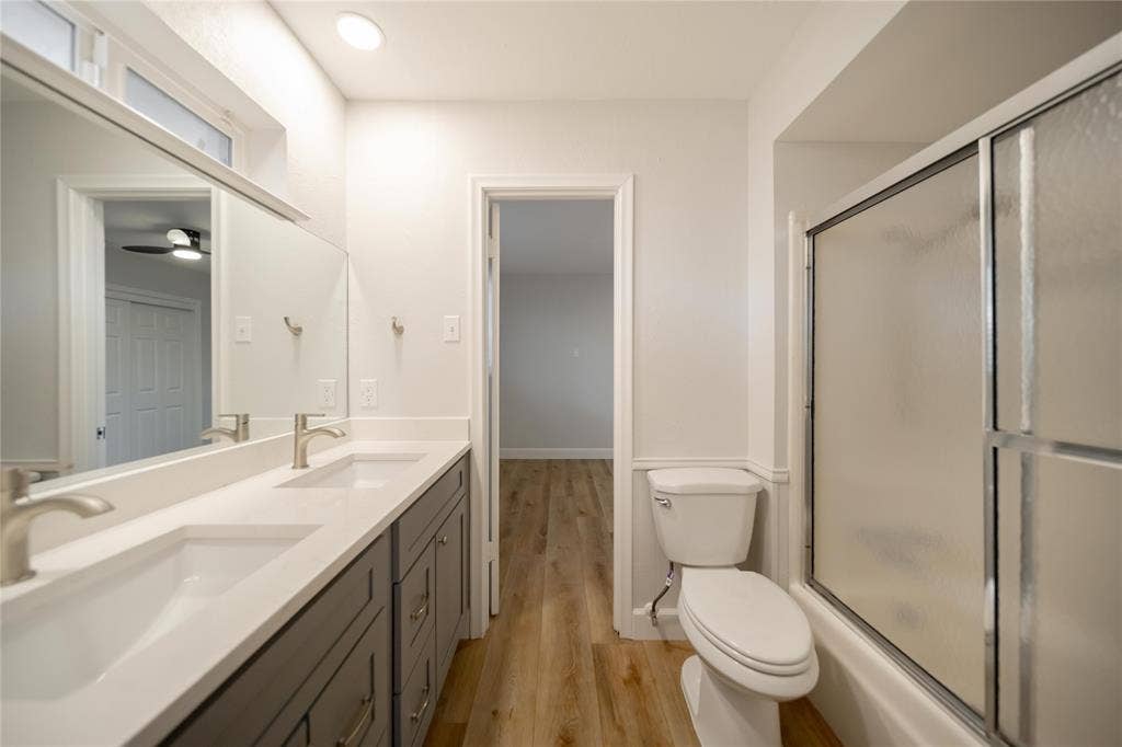 4333 Harvest Hill Road, Dallas, TX 75244 Dual-sink vanity with white countertops and gray cabinetry, large wall-mounted mirror, wood-finish flooring, and a shower-tub combination with sliding glass doors