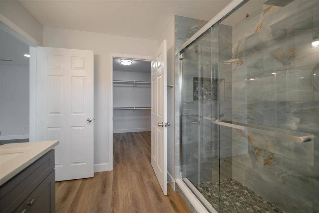 4333 Harvest Hill Road, Dallas, TX 75244 Walk-in shower featuring large-format tile walls, integrated shelving niche, and pebble-style floor tiling, complemented by a wood-finish vanity and walk-in closet with built-in shelving