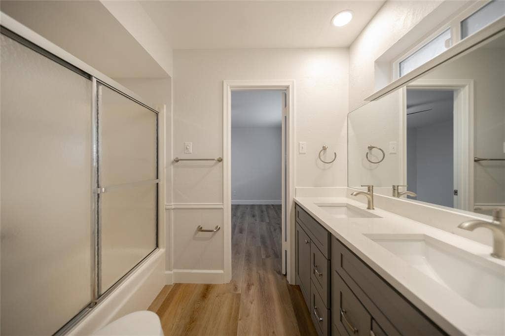 4333 Harvest Hill Road, Dallas, TX 75244 Bathroom featuring a double vanity with grey cabinetry, a solid surface countertop with integrated dual sinks, a full-width vanity mirror, wood-finish flooring, and a tub-shower combination with a sliding glass enclosure