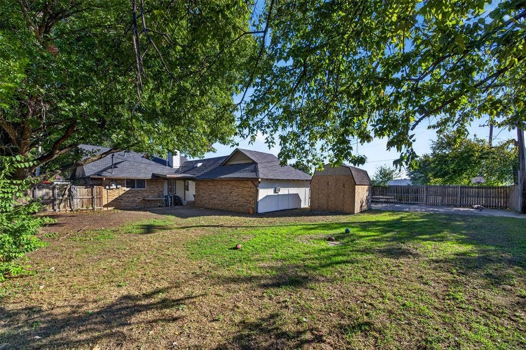 113 Dove Avenue, Red Oak, TX 75154 Spacious backyard featuring brick exterior siding, a shingled roof, mature tree canopy, and an attached storage structure enclosed by perimeter wood fencing