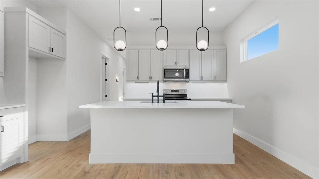 2816 Marburg Street, Dallas, TX 75215 Kitchen featuring a central island with a solid-surface countertop, shaker-style cabinetry, stainless steel appliances, matte black plumbing fixtures, and three glass pendant lights