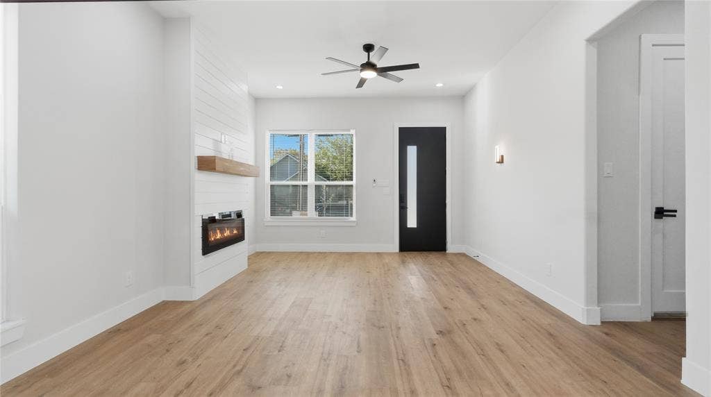 2816 Marburg Street, Dallas, TX 75215 Living area featuring wood-finish flooring, a wall-mounted electric fireplace with a floating wood mantel, horizontal shiplap detailing, a contemporary ceiling fan, and a solid door with a vertical glass pane