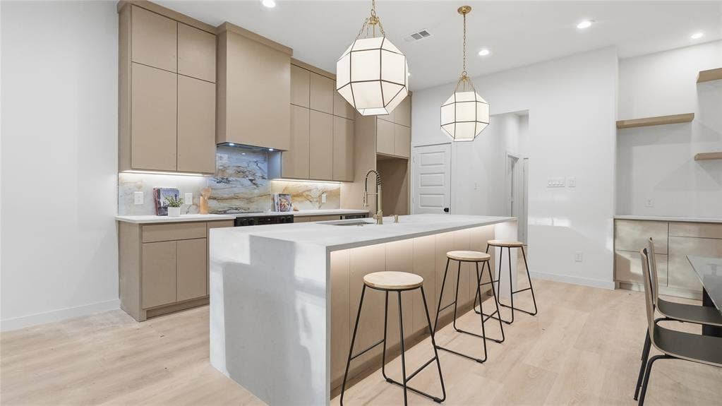 Modern kitchen featuring flat-panel cabinetry, a waterfall-edge center island with integrated task lighting, stone-patterned backsplash, and wood-finish flooring