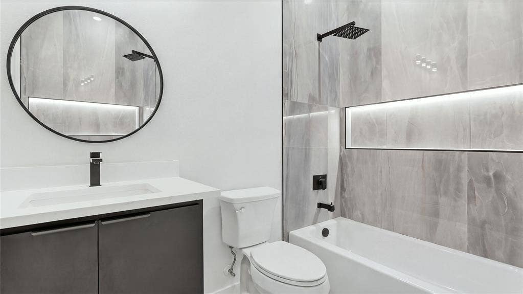 Modern bathroom featuring a dark vanity with a white countertop, circular mirror, matte black fixtures, and large-format wall tile with integrated recessed lighting