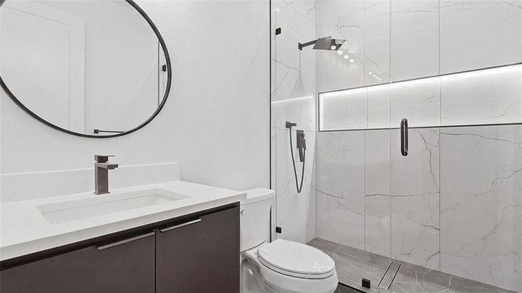 Bathroom featuring a floating dark-finish vanity with a rectangular sink, wall-mounted matte black faucet, oversized circular mirror, glass-enclosed shower with large-format tile surround, and built-in recessed lighting